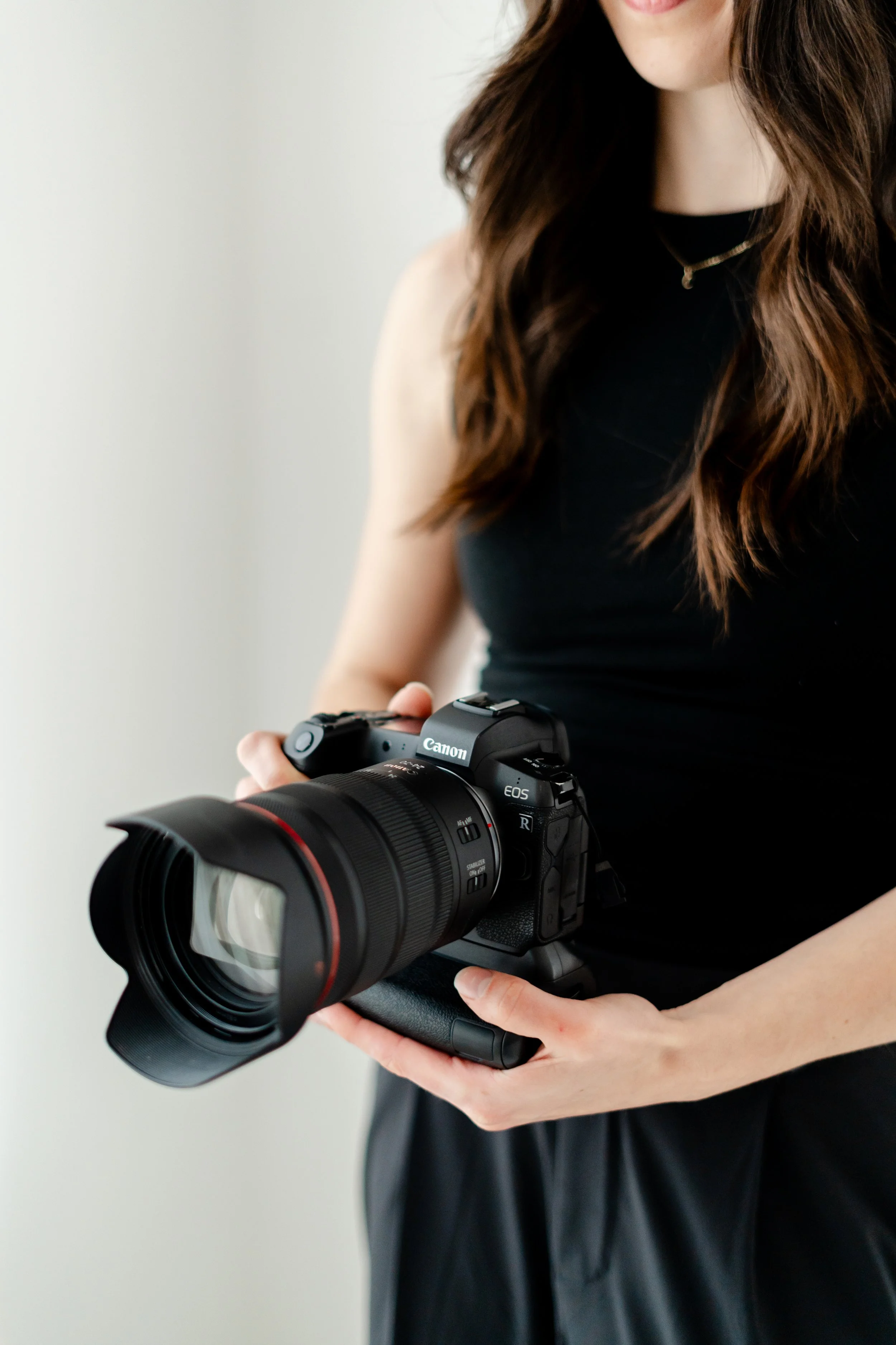 A person holding a Canon EOS R camera with a large lens, dressed in black top and skirt, with brown wavy hair, against a plain background.