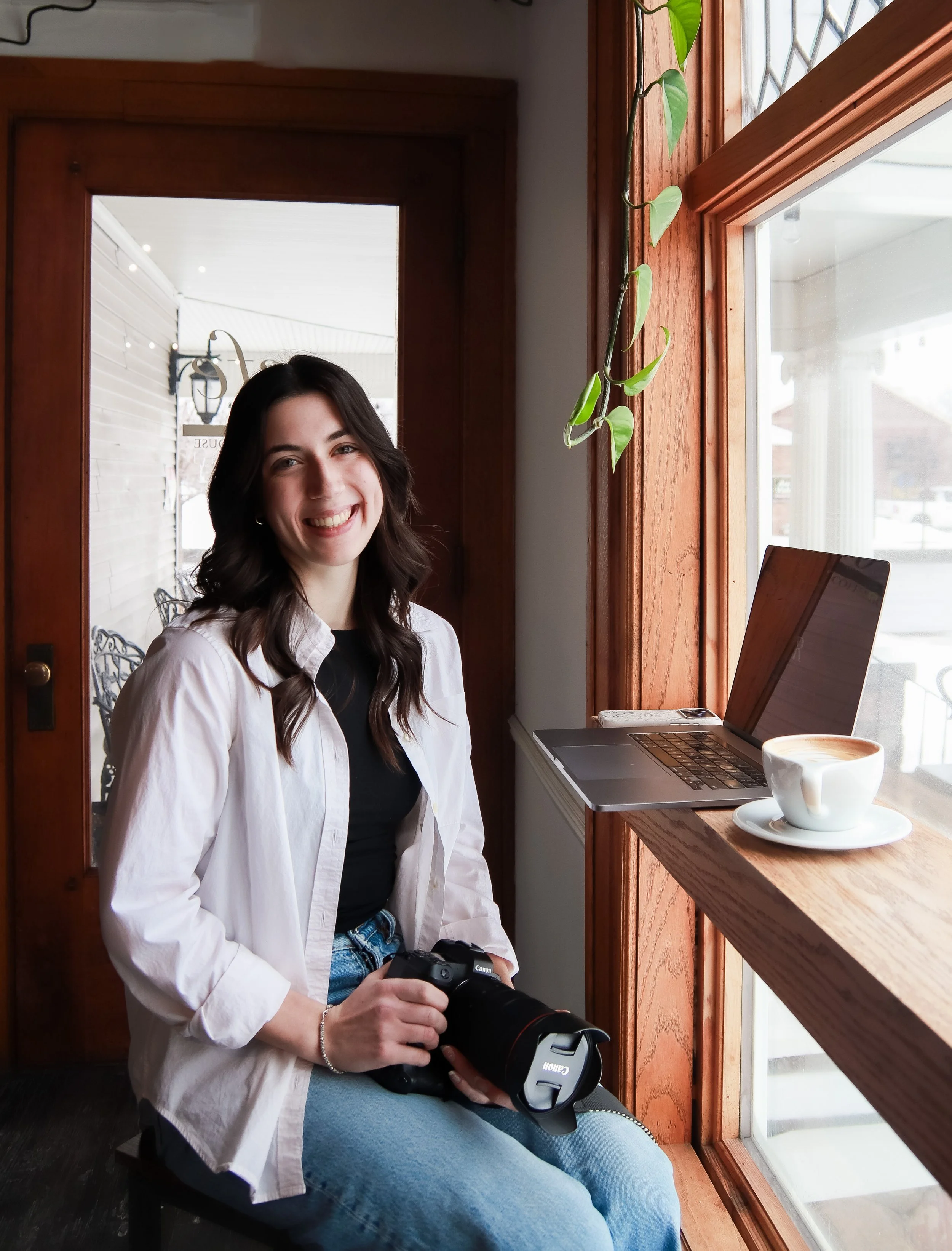 A young woman (Michaela Mueller, Founder of ROVA) sitting by a large window in a cafe, smiling, holding a DSLR camera in her lap. There is a laptop and a cup of coffee on the windowsill beside her, and a green plant hanging from the window frame.