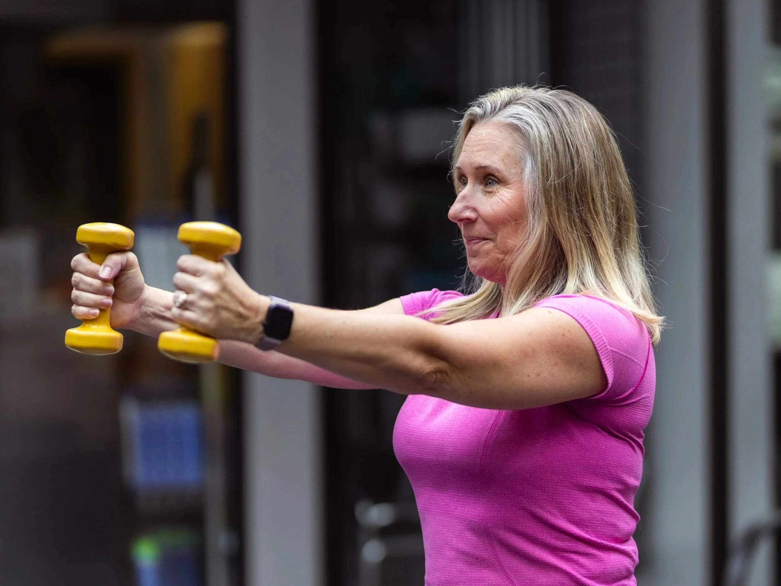 An elderly woman in a pink shirt lifting yellow dumbbells in a gym.