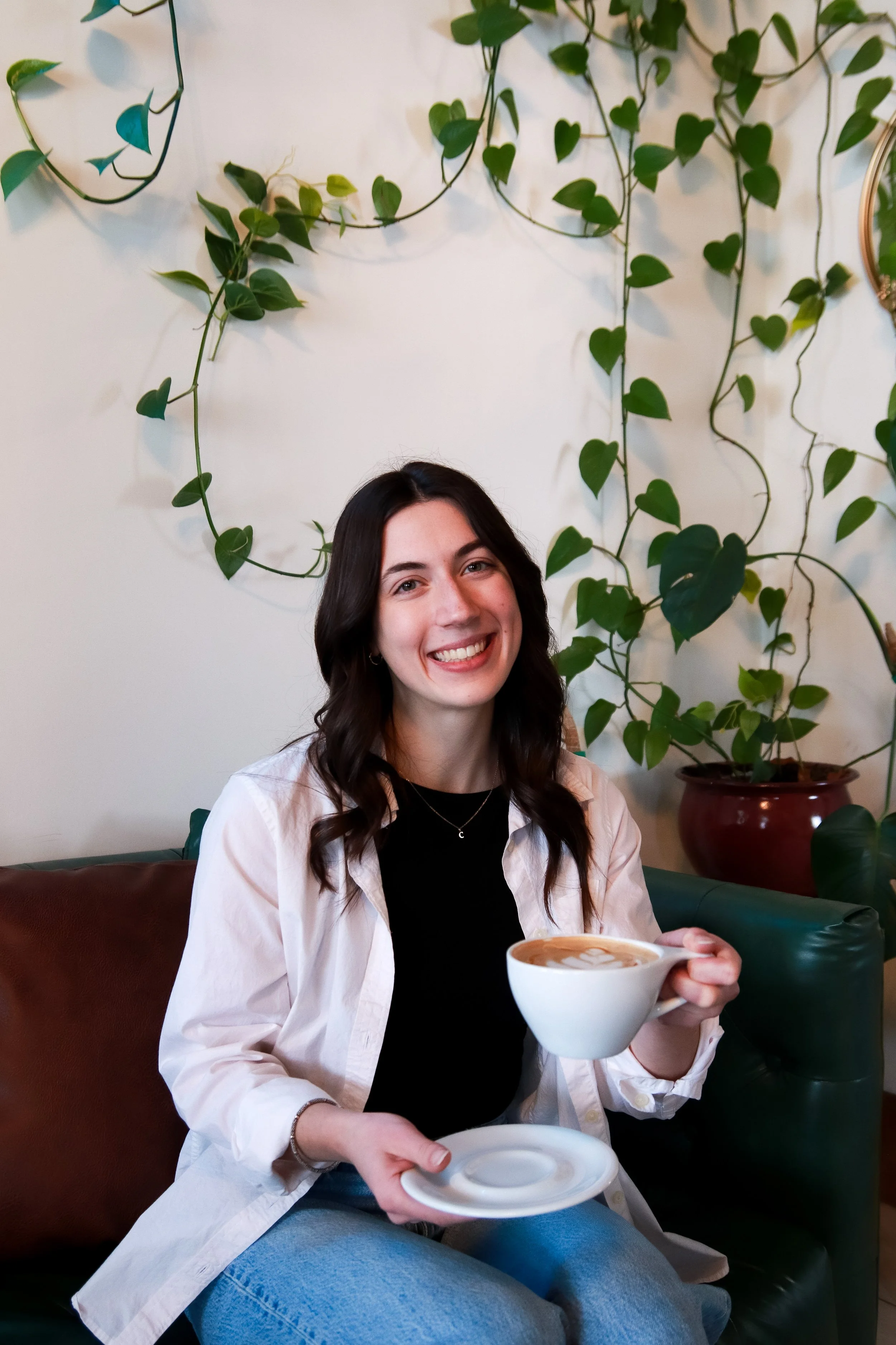 A smiling woman (Michaela Mueller, Founder of ROVA) with dark wavy hair sitting on a green sofa holding a cup of coffee or latte and a saucer in a cozy café decorated with green potted plants and a white wall.
