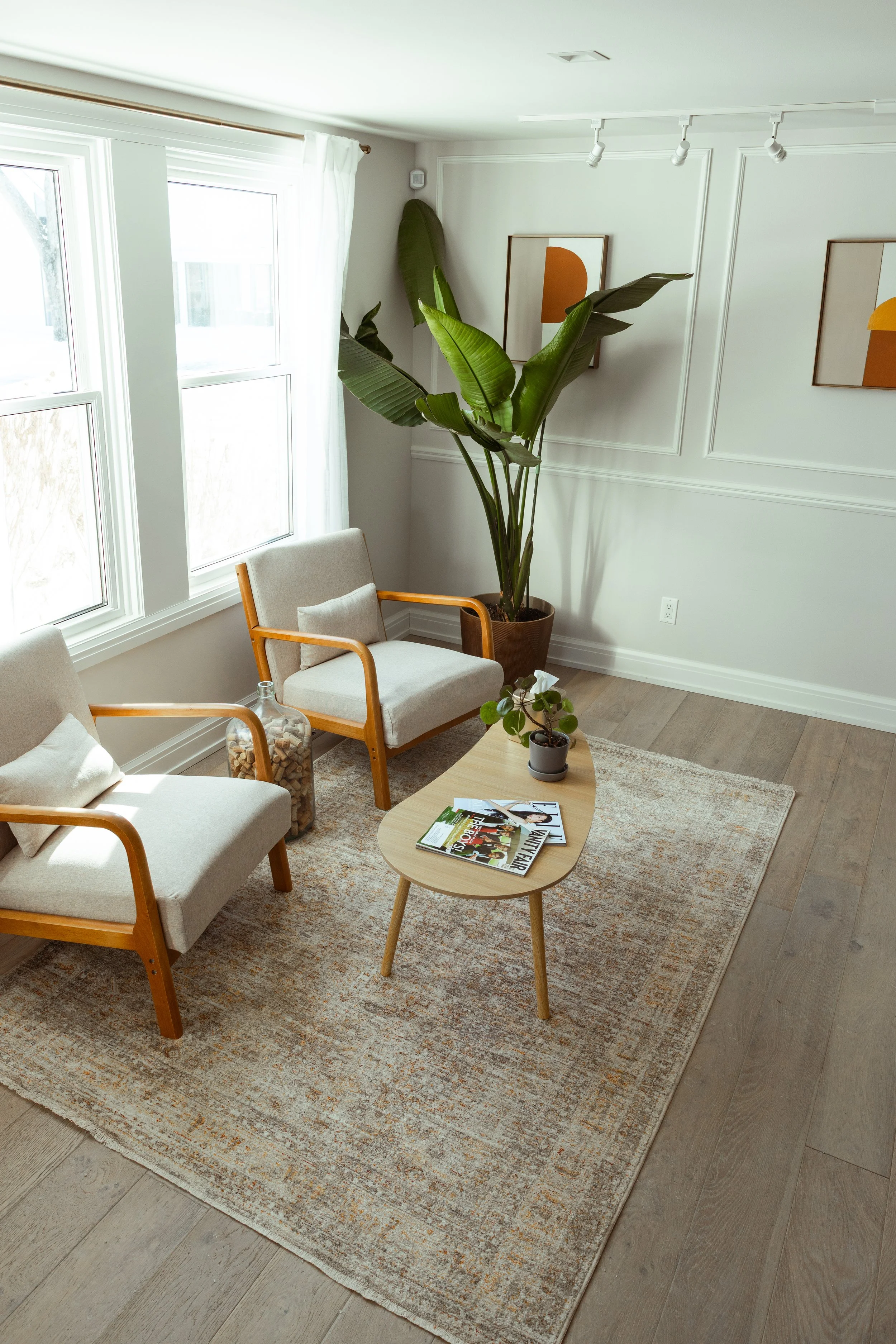 A cozy living room corner with two beige armchairs with wooden arms, a small oval wooden coffee table with magazines and potted plants, a large potted plant with broad leaves, two framed abstract art pieces on the wall, a window with white curtains, a beige area rug, and light wood flooring.