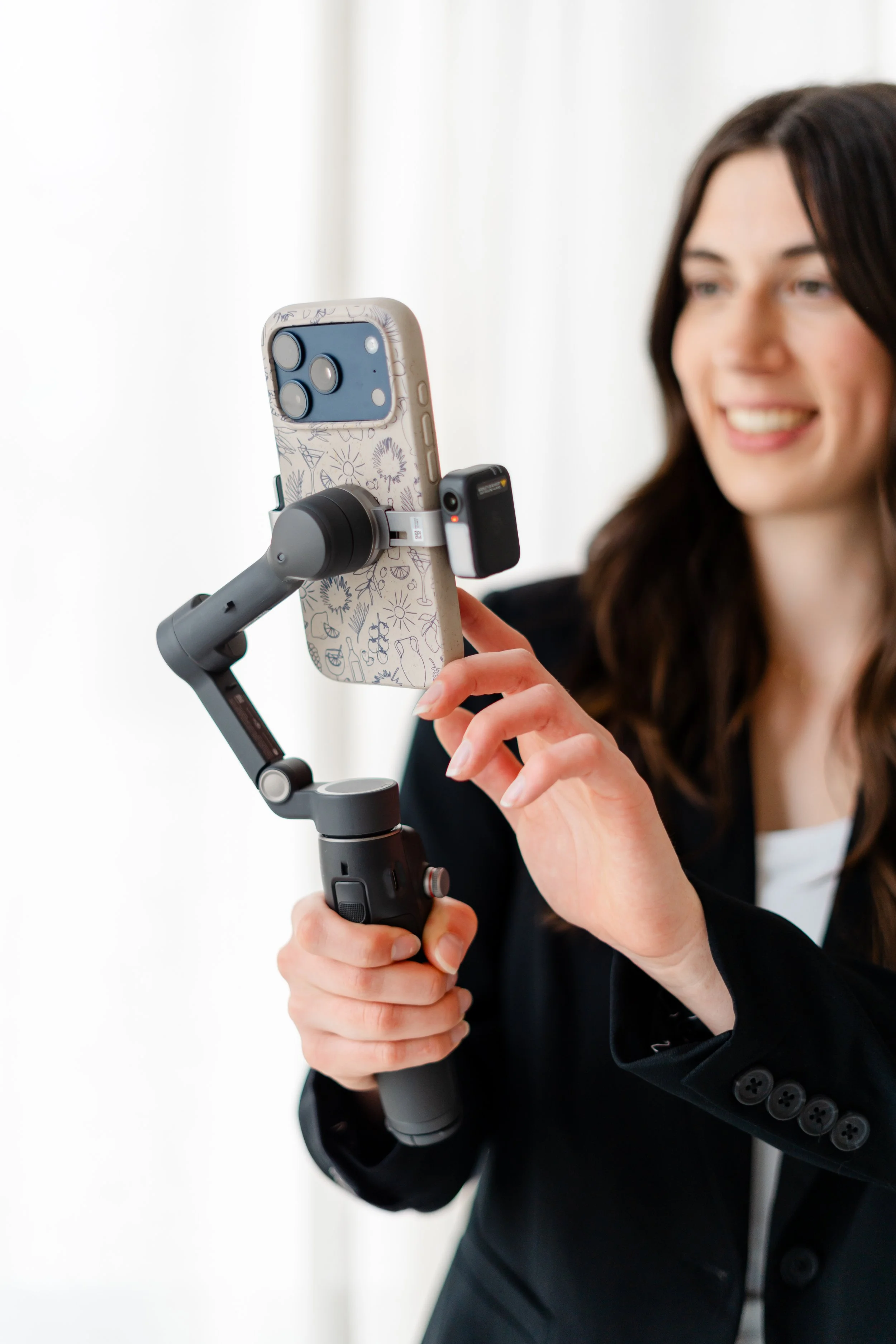 A woman with long dark hair in a black blazer is smiling and taking a selfie with a smartphone mounted on a stabilizer.