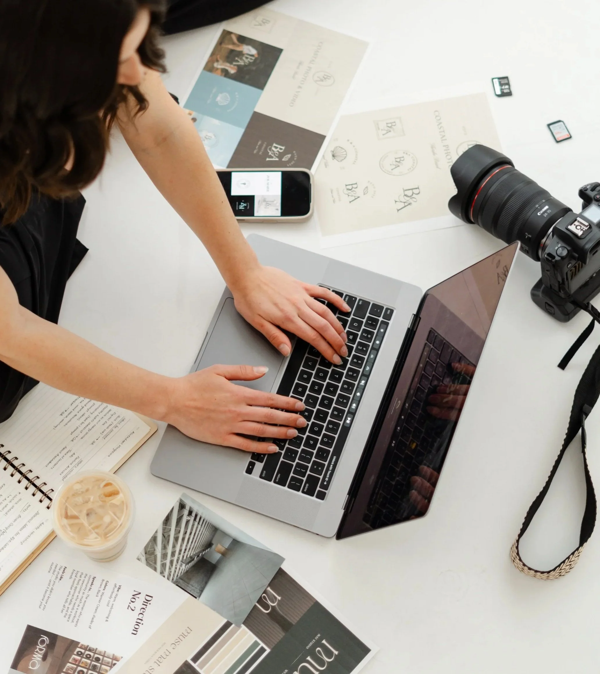 A person working on a laptop with photography equipment and printed materials on a white table, including business cards, brochures, and a beverage in a plastic cup.