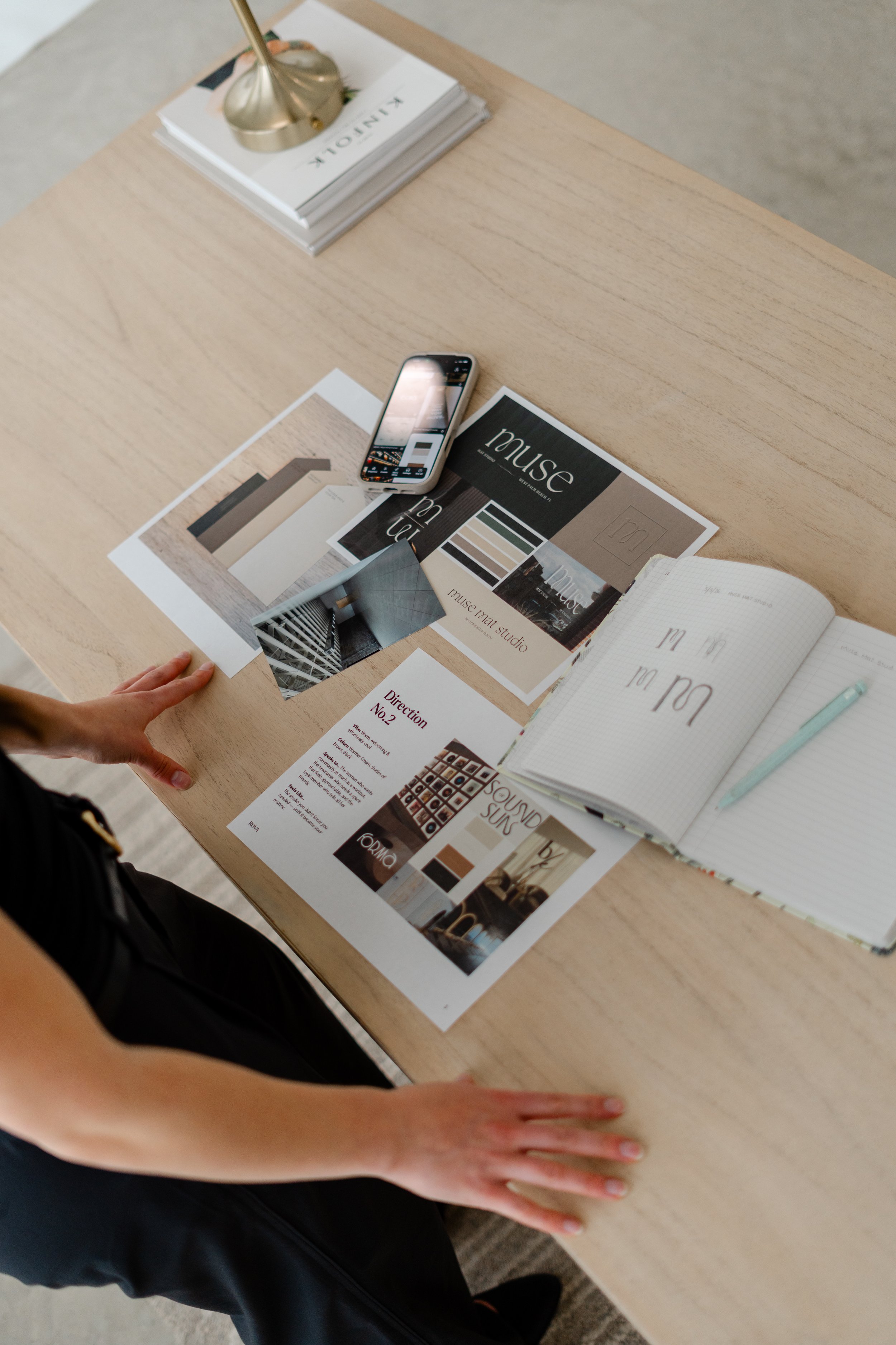 A person with a black sleeveless top is leaning over a light wood table. On the table, there are magazines, paper samples, and a smartphone. The person's left hand is resting on the table, and their left hand is slightly visible.