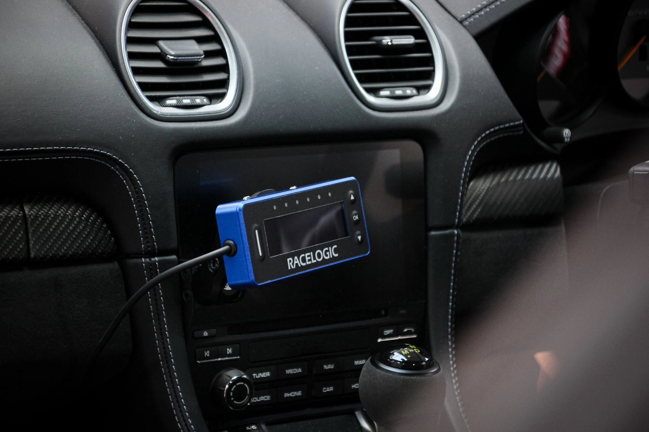 Close-up of a car's dashboard, showing the centre console with air vents, a blue device labelled 'RACE LOGIC' connected by a wire, and various control buttons.