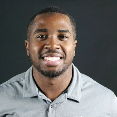 A smiling man with short hair and a beard, wearing a gray collared shirt, against a dark background.