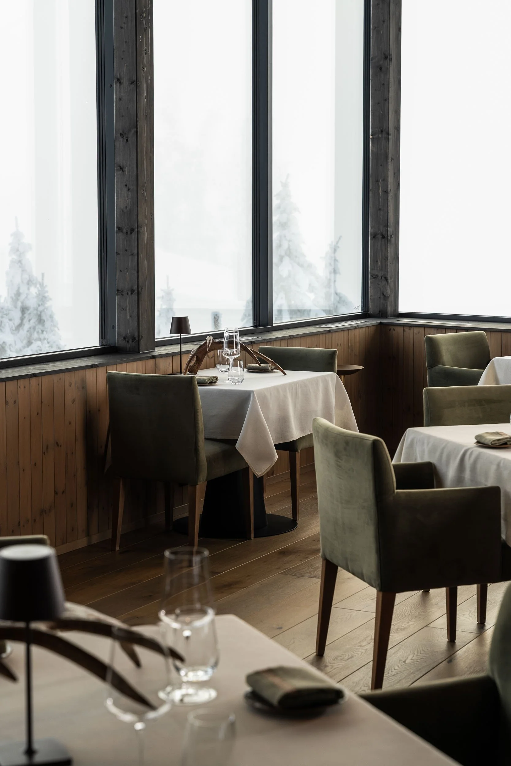 Interior of a restaurant with tables set with glasses, napkins, and plates. Large windows with black frames show snowy trees outside. The walls are wooden-paneled, and there are green upholstered chairs around the tables. A small black table lamp is on one of the tables.