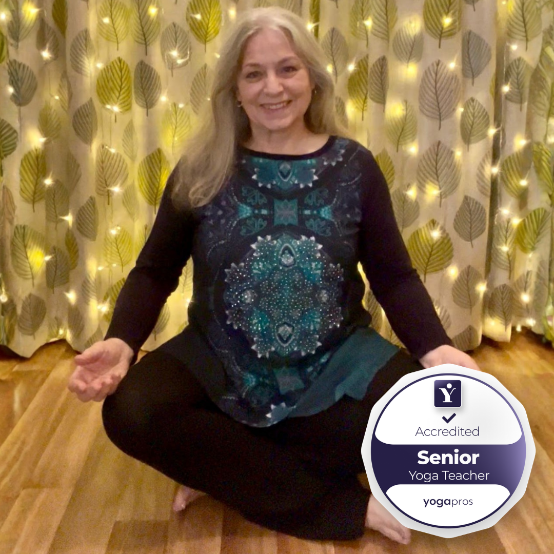 A smiling woman sitting cross-legged on a red, patterned yoga mat surrounded by colorful singing bowls, lit candles, small pink and white candles, a bundle of lavender, and a water bottle, in a room with chairs against the wall. There is a logo in the bottom right corner that reads 'Yoga Alliance Professionals Senior Yoga Teacher'.
