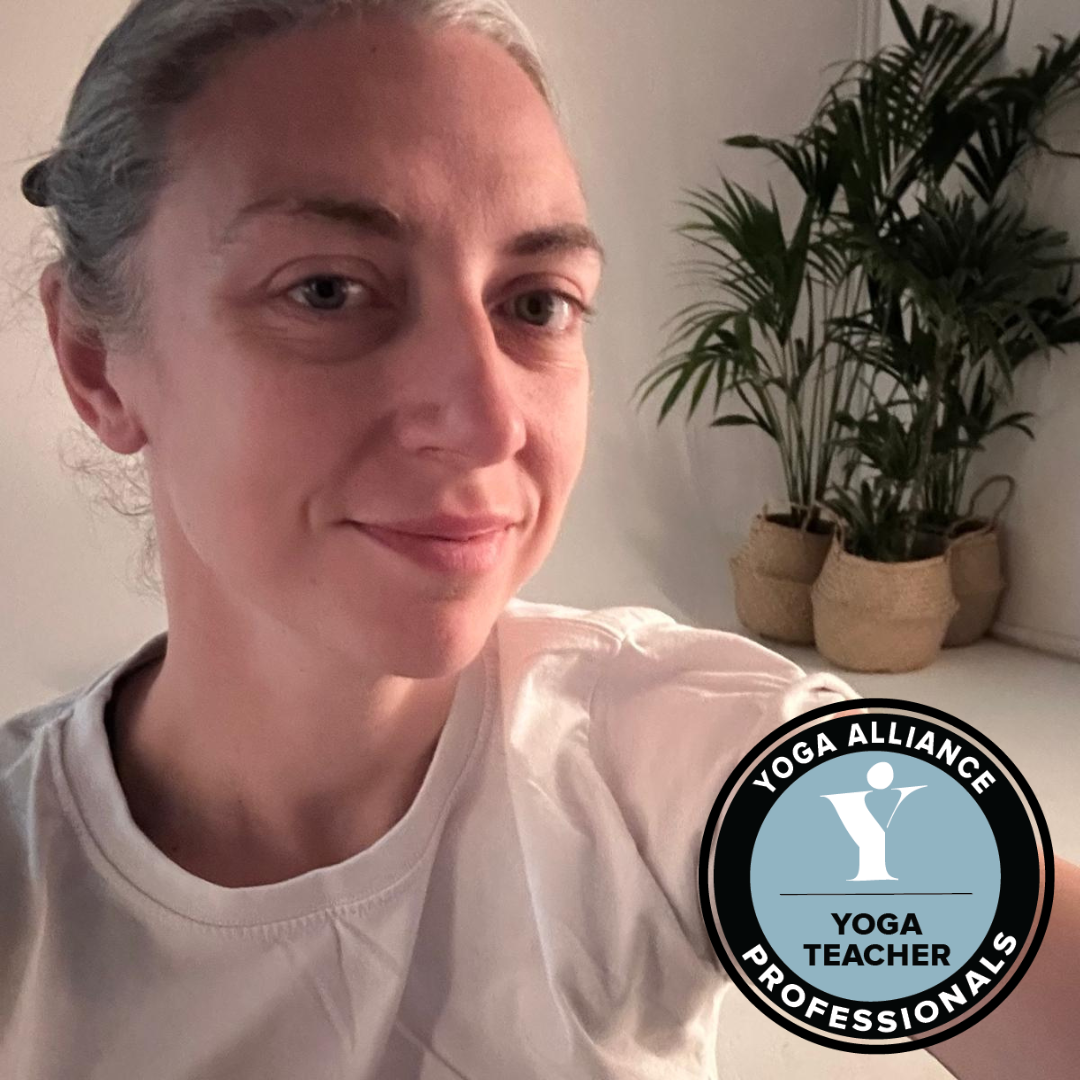 A smiling woman with short gray hair, dressed in a purple sleeveless top, sitting against a black background. She has tattoos on her arm and is associated with the Yoga Alliance as a yoga teacher professional.