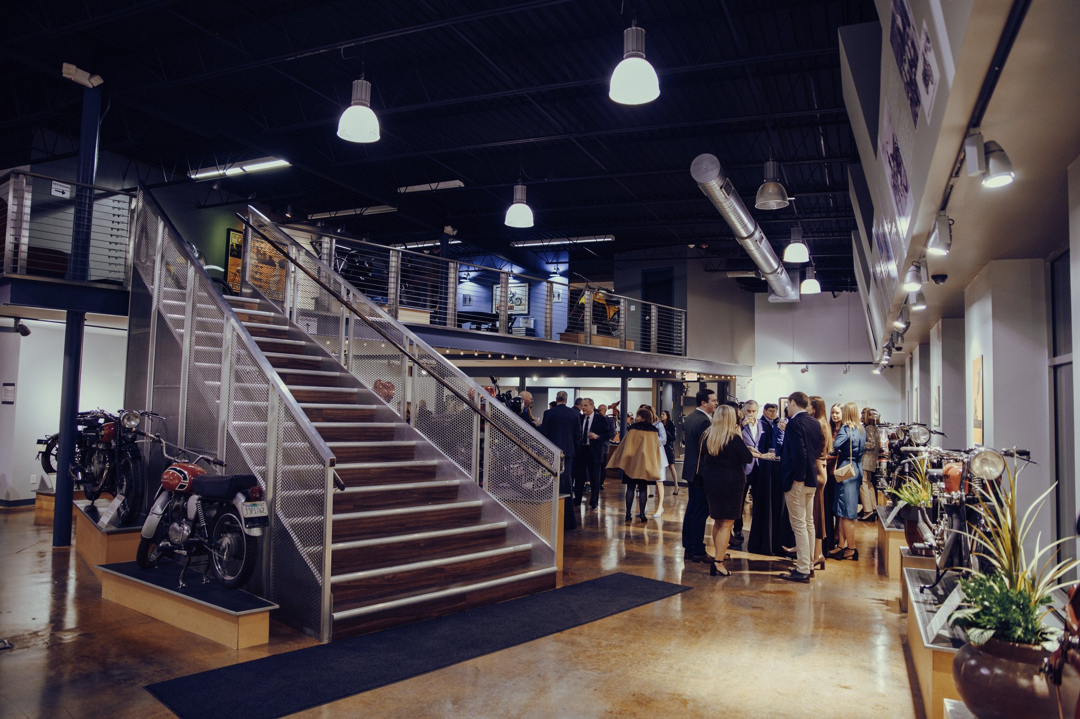 view of interior east gallery at the moto museum event venue st. louis. crowd is standing during cocktail reception and a set of stairs leads to mezzanine motorcycle gallery