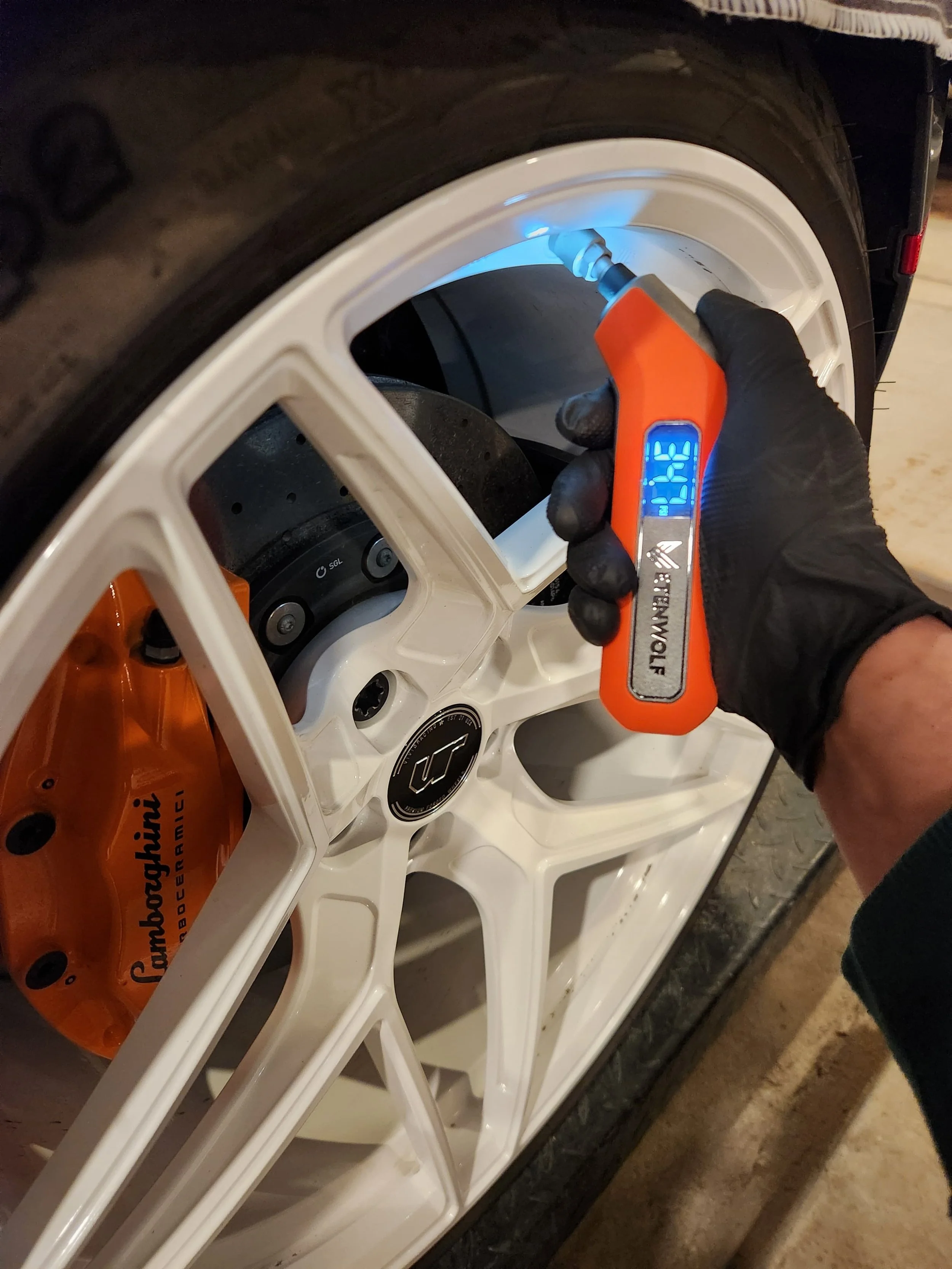 A person in black gloves using a handheld infrared thermometer to measure the temperature of a white Lamborghini wheel.