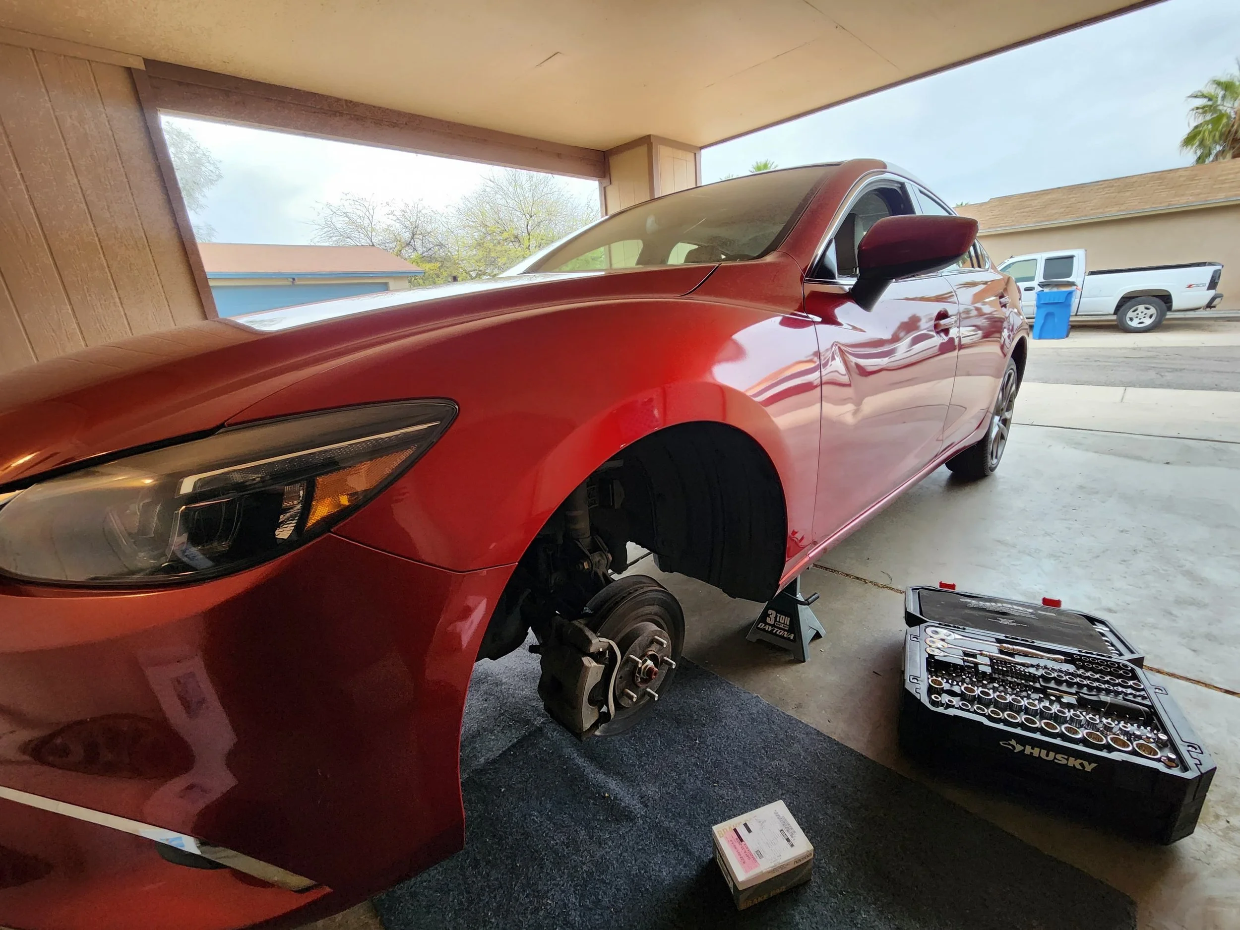 Red car with front wheel removed, supported by a jack stand in a garage, with a toolbox and small parts on the floor.