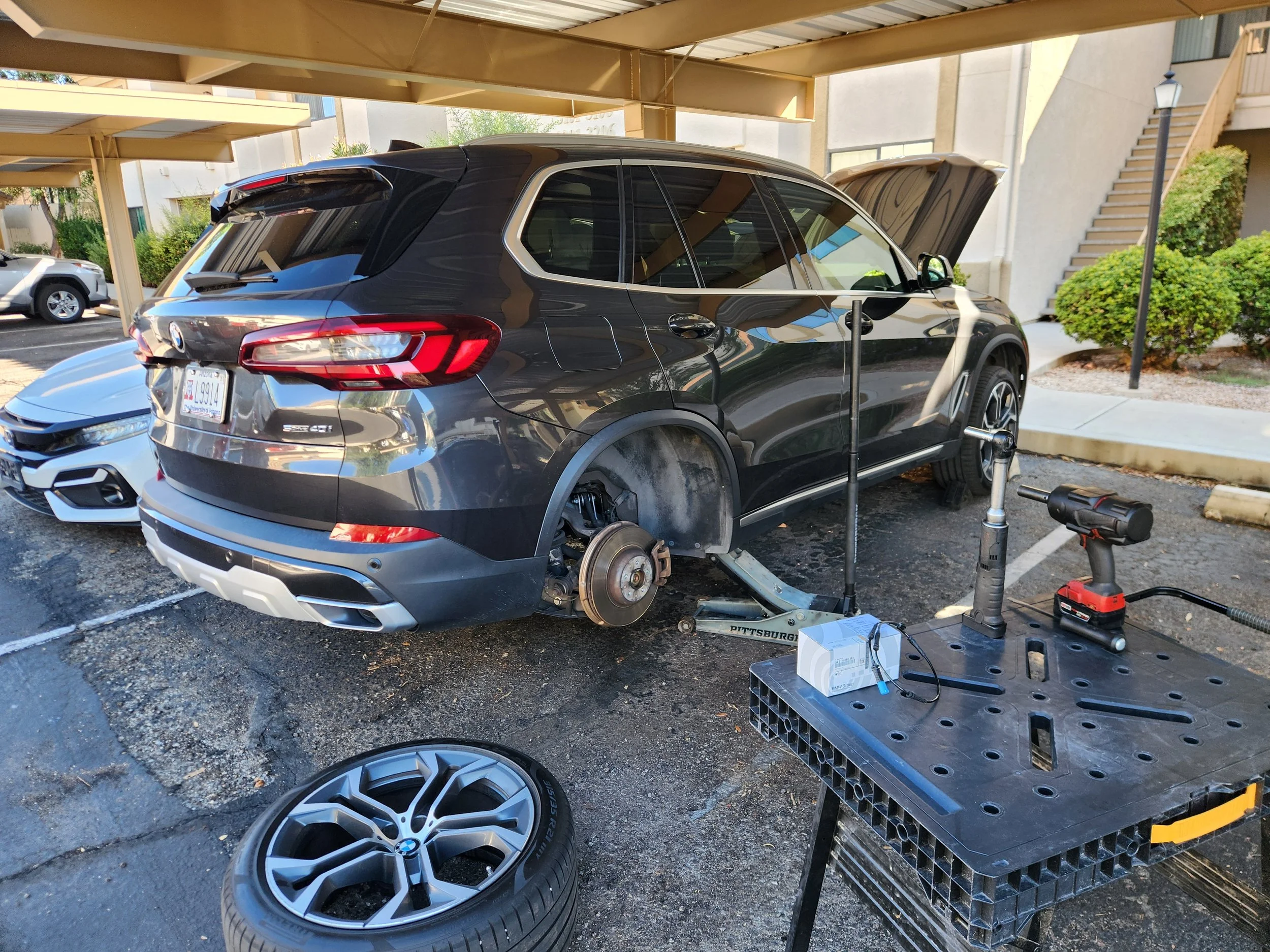 A black BMW SUV is raised on a hydraulic lift with its rear wheel removed, in an outdoor parking area under a carport. An extra BMW wheel with tire is on the ground nearby. A cordless drill, a work light, and other tools are on a black workbench.