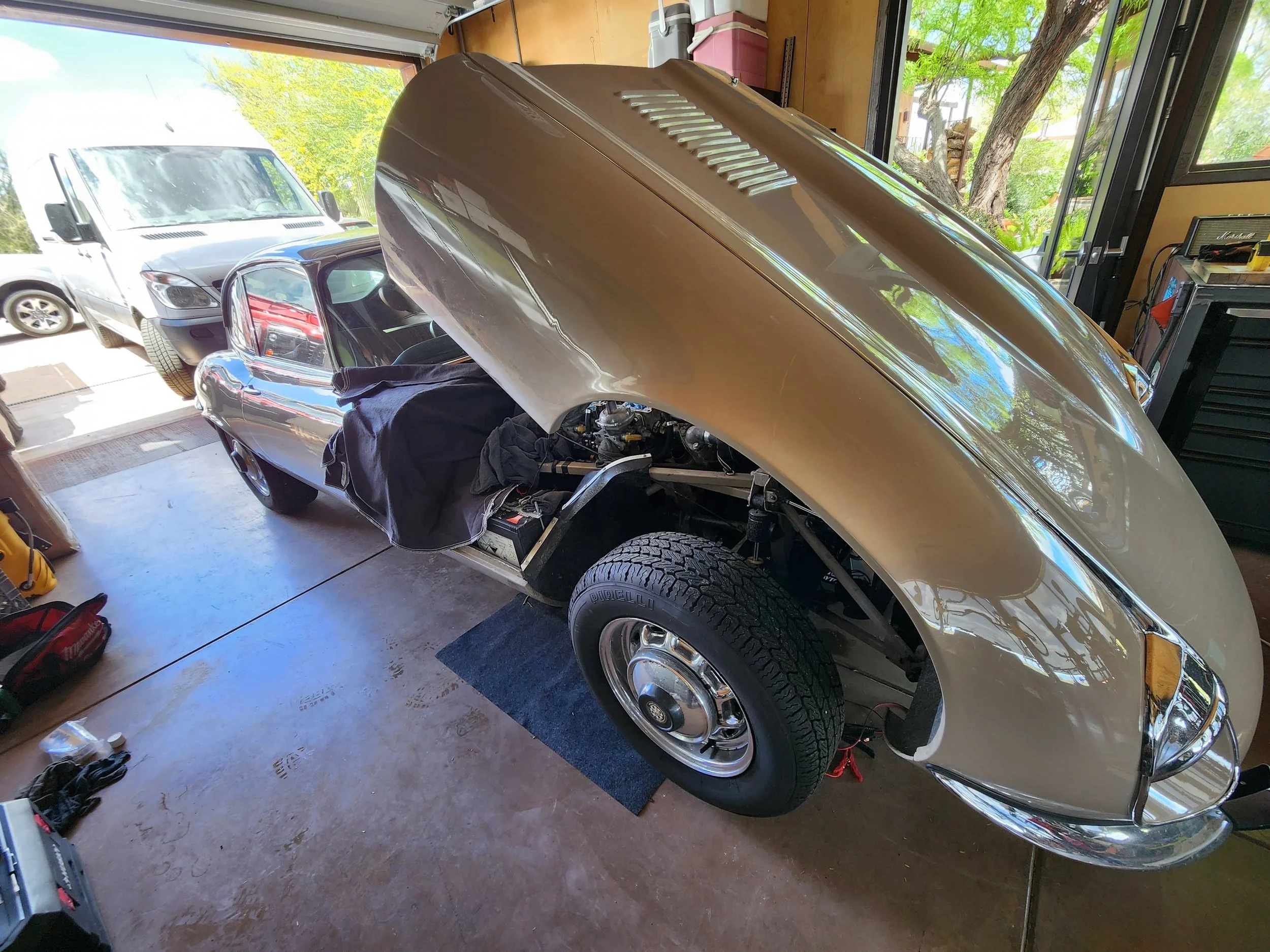 A classic car with its front hood open, situated inside a garage. The garage has tools and a workbench, and a large window shows trees and parked cars outside.
