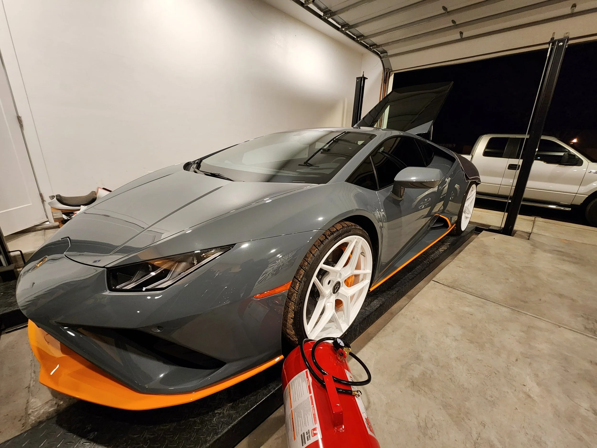 Gray sports car with white wheels inside a garage, with a fire extinguisher in front of it and a pickup truck behind.
