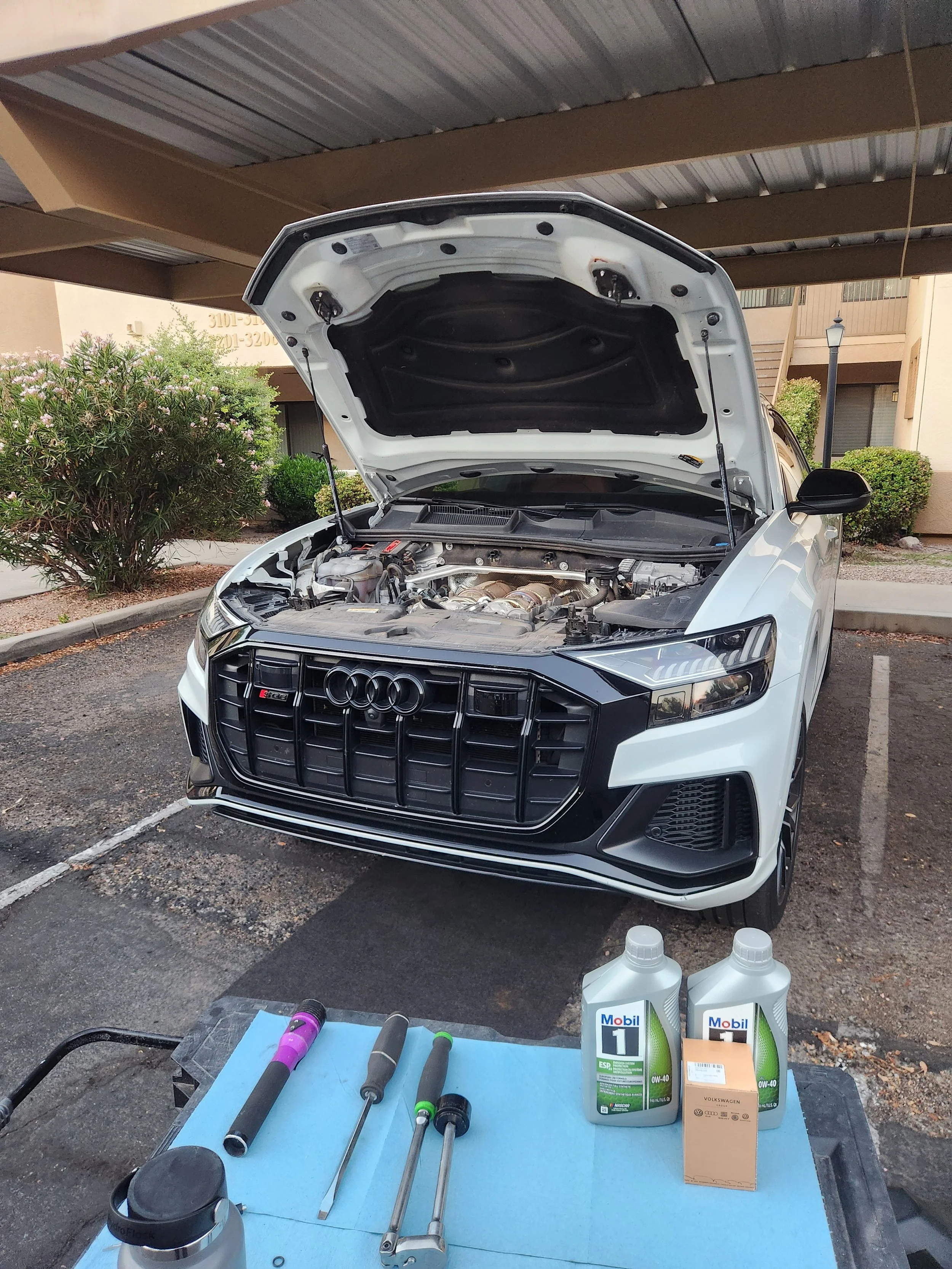 A white Audi car with its hood open parked in a parking lot. There are tools and bottles of motor oil on a table in front of the car, indicating an oil change or vehicle maintenance.