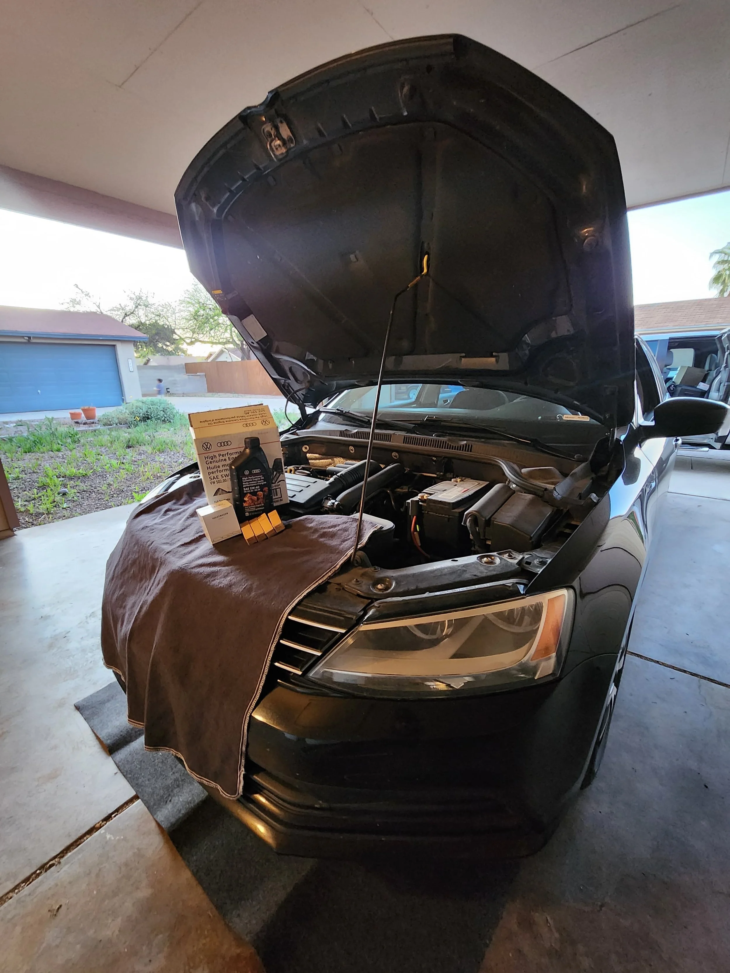 A black sedan with its hood open, parked in a garage, with a brown towel draped over the front fender and automotive products placed on top.