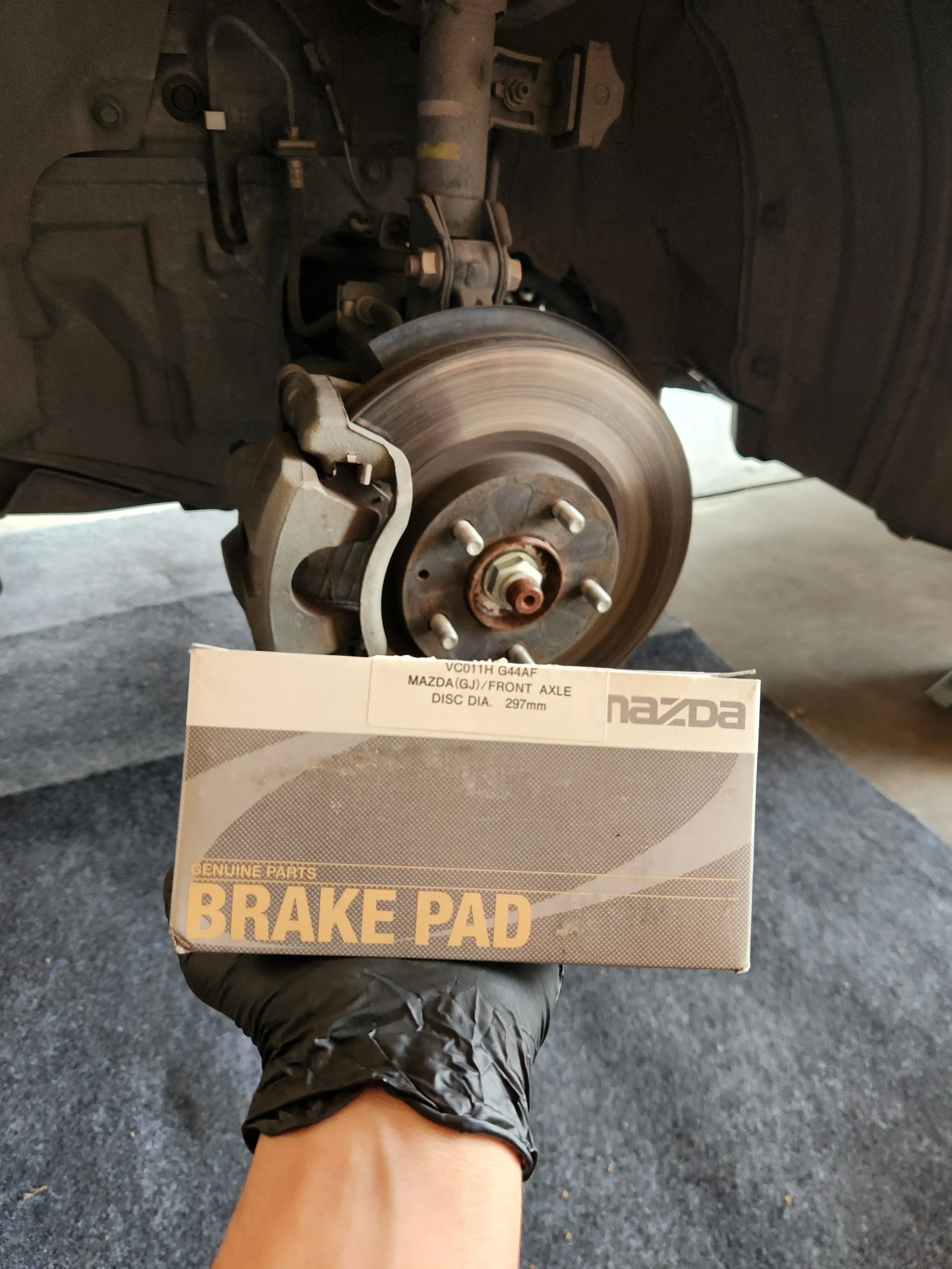 A person holding a Mazda brake pad in front of a car's front wheel assembly, which is partially disassembled showing the brake rotor and caliper.