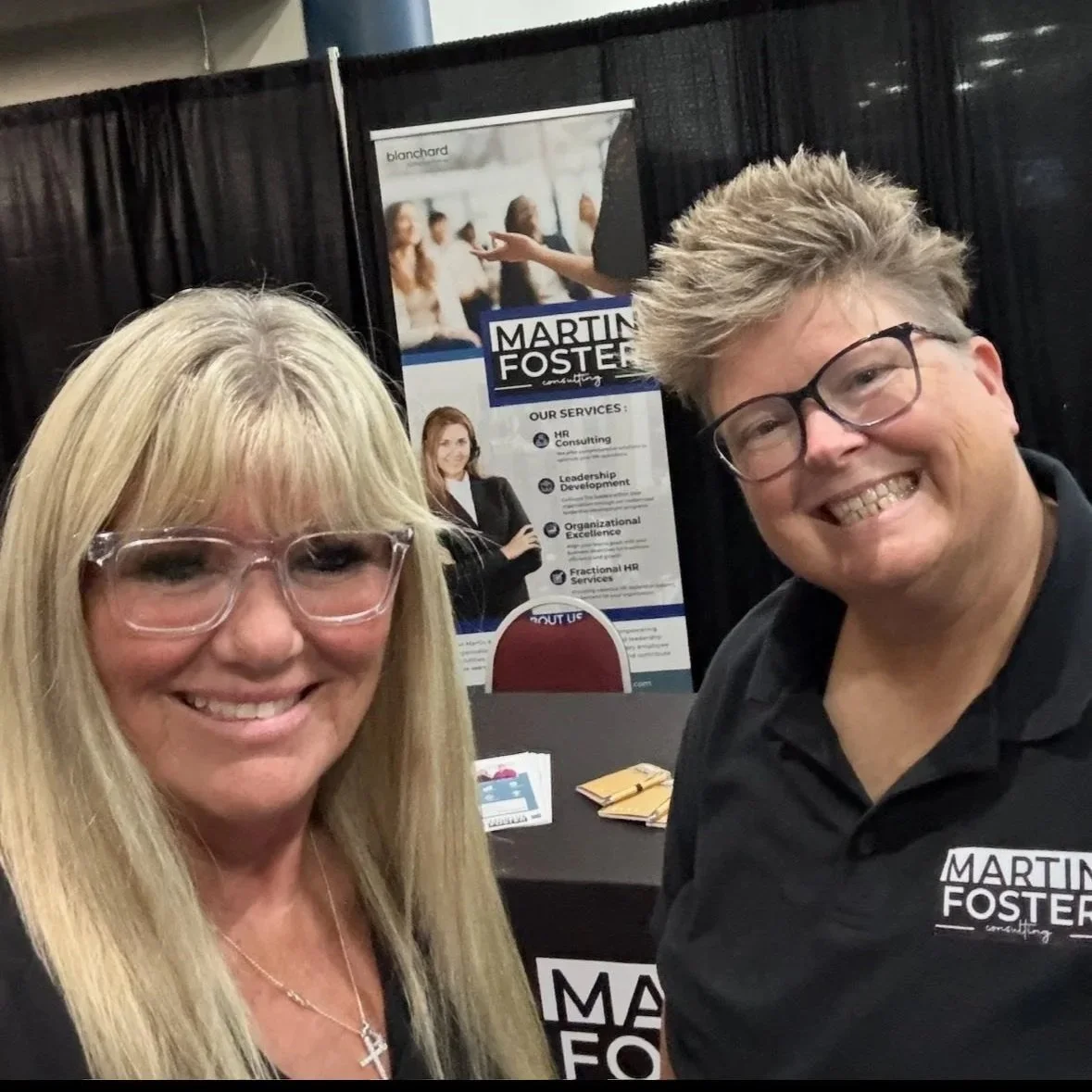 Anita Martin and Shannon Foster of Martin and Foster Consulting smile at a public event booth, representing their approachable, expert-led HR brand. Anita wears glasses and has long blonde hair; Shannon, also wearing glasses, has short spiky hair