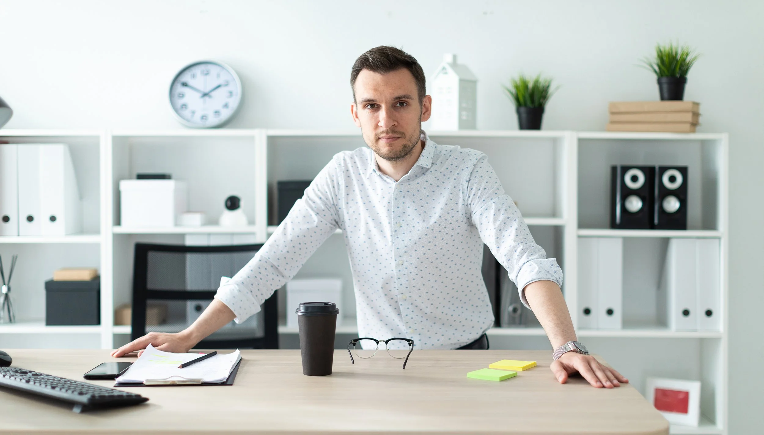A man stands confidently in a clean, organized office space, surrounded by bookshelves, plants, and workspace essentials — reflecting clarity, professionalism, and readiness to help.