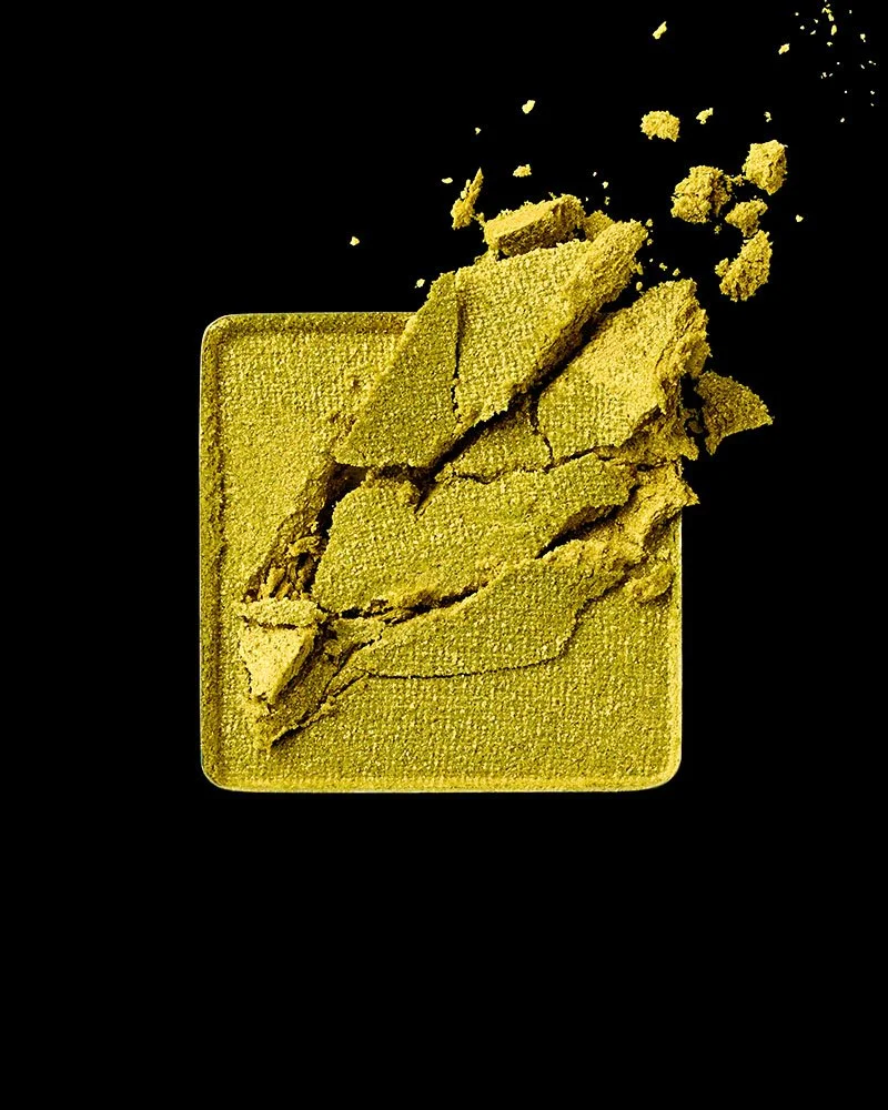 A broken compact of green eyeshadow with powder scattered on a black background.