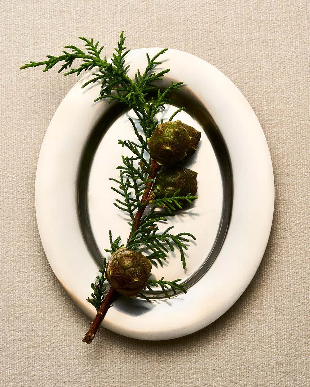 A white oval plate with a black and white decorative border, holding a sprig of green evergreen with three pine cones, placed on a beige textured surface.