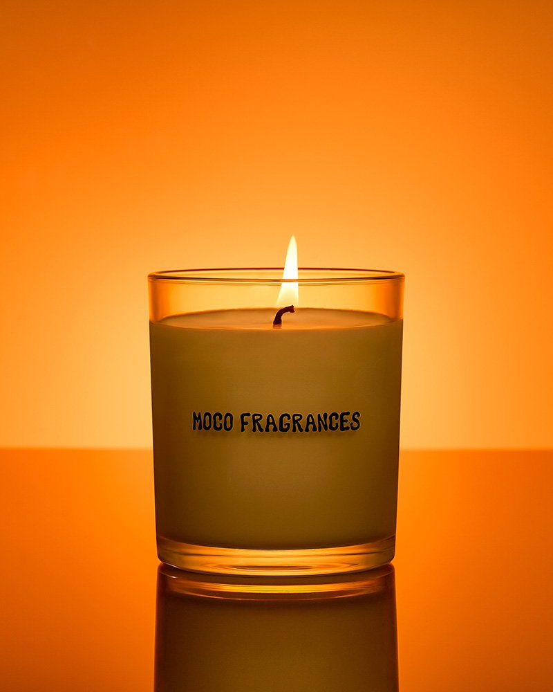 A lit candle in a glass container with the text 'MOCO FRAGRANCES' on it, against an orange background.