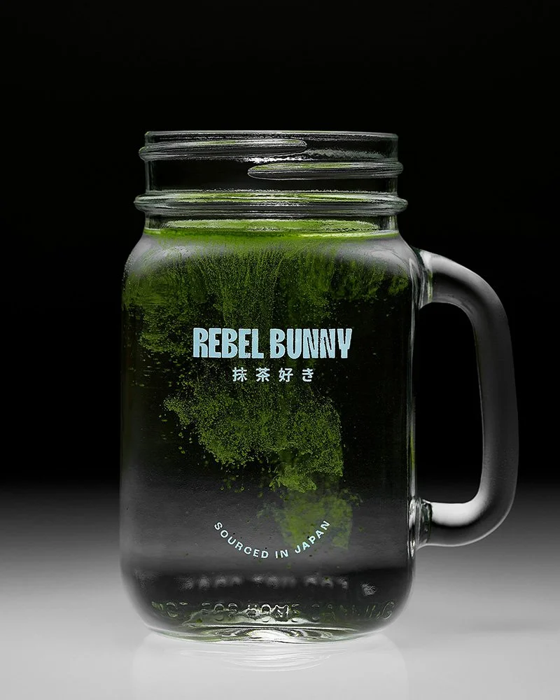 A clear glass mug with a handle filled with a green liquid and green powder inside, labeled 'Rebel Bunny' with Japanese characters and 'Sourced in Japan' on a black background.