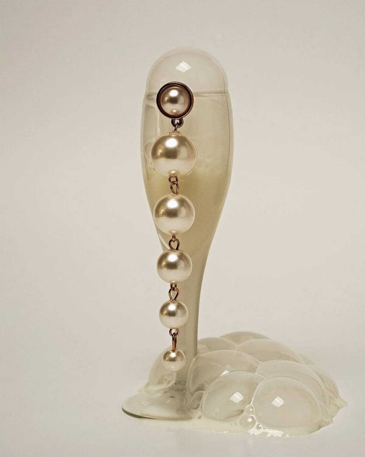 Dangling strand of pearl earrings hangs off an apertif glass of milk bubbles on a white display surface. Editorial still-life jewelry photography by photographer Sara Anderson.