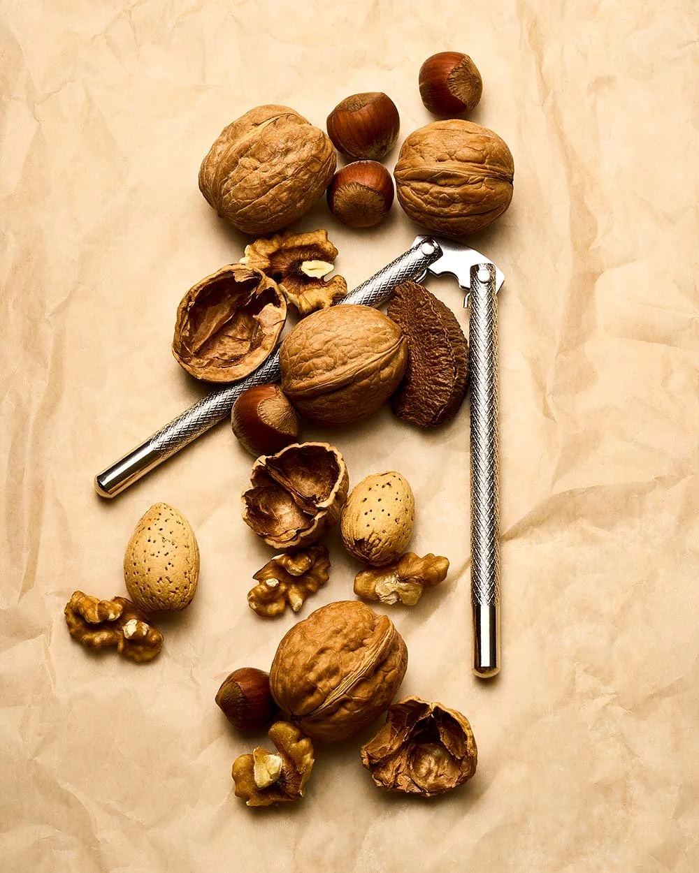 A collection of walnuts, hazelnuts, almonds, and two nutcrackers arranged on a crumpled piece of paper.