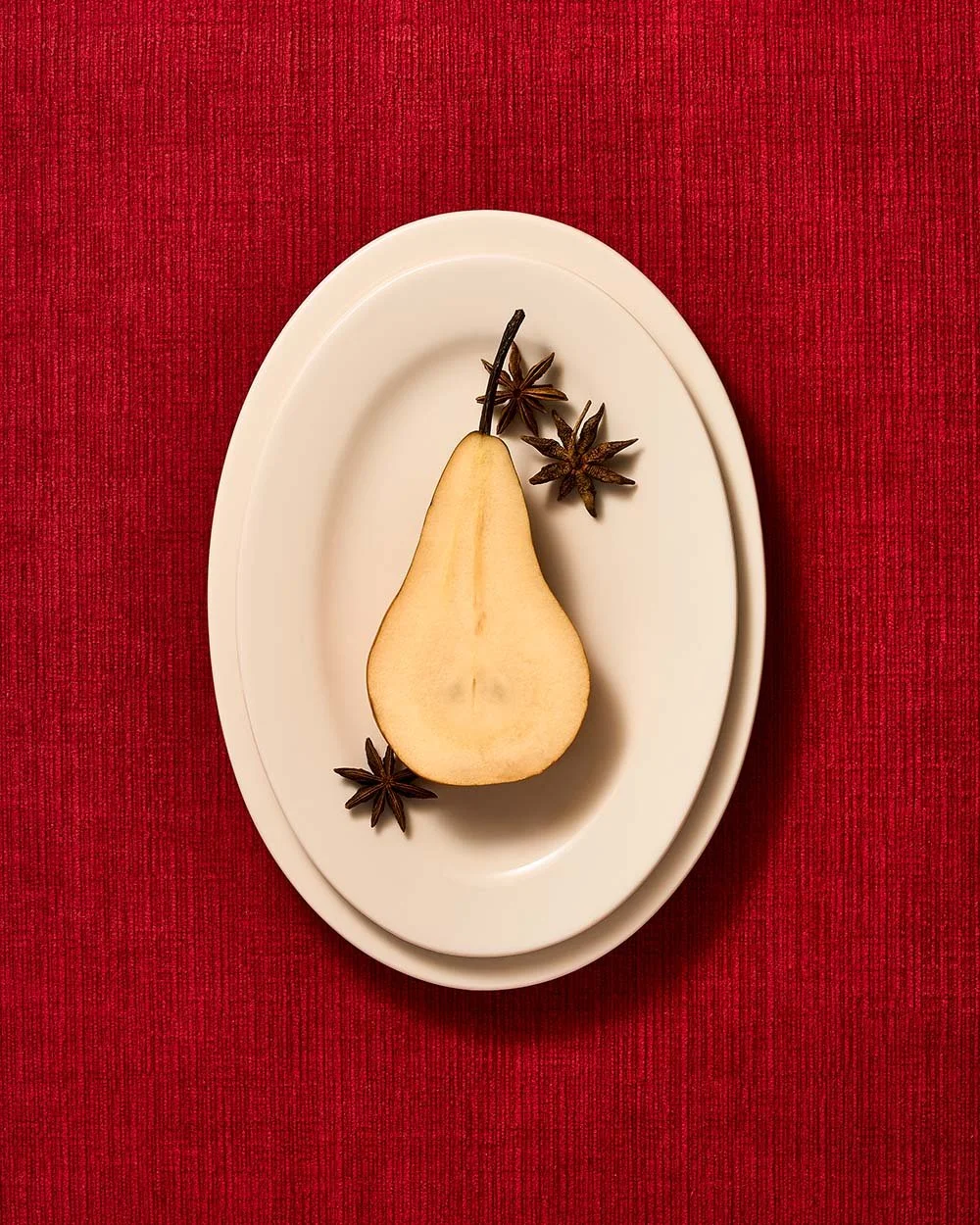 A white pear and three star anise pods on a white oval plate placed on a red textured tablecloth.