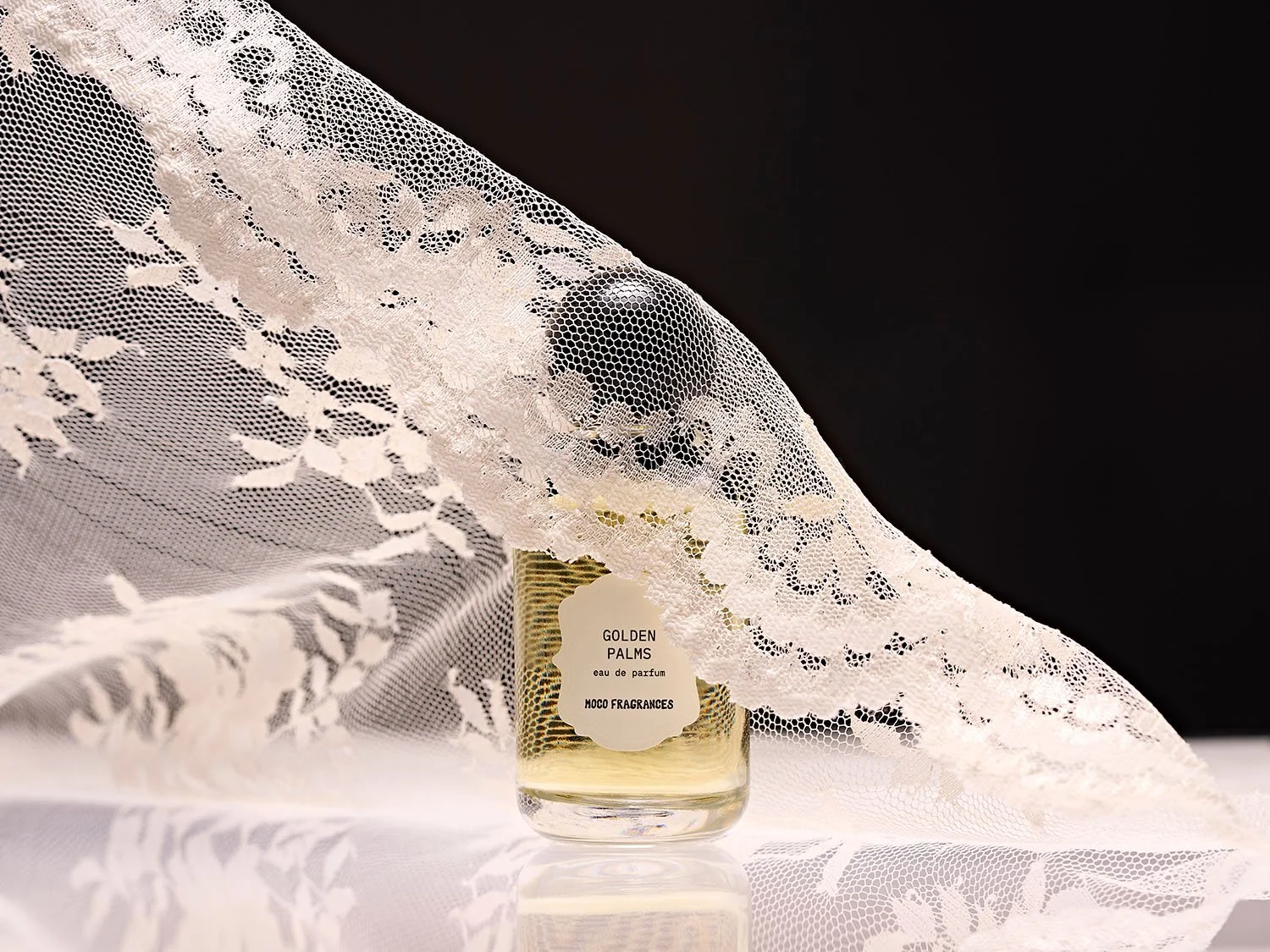 MOCO Golden Palms perfume bottle partially concealed beneath draped white floral lace against a black background. Editorial fragrance photography by photographer Sara Anderson.