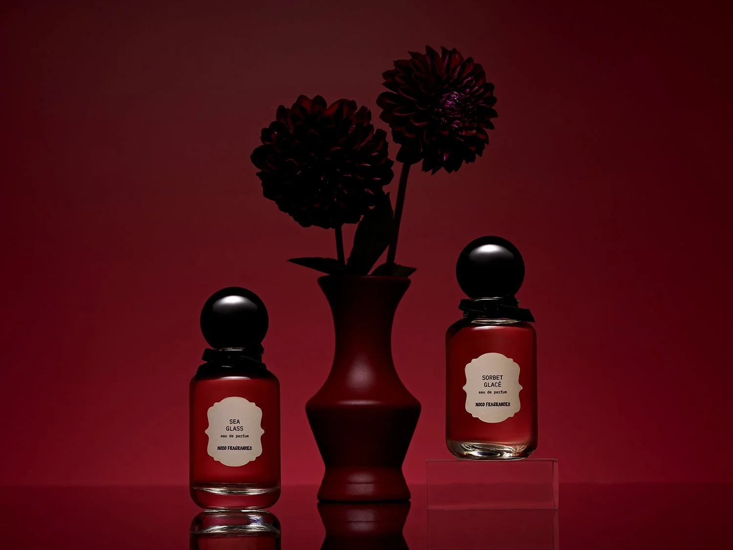 MOCO Sea Glass and Sorbet Glacé perfume bottles arranged around a red vase of dark dahlias against a deep red background. Commercial fragrance photography by photographer Sara Anderson.