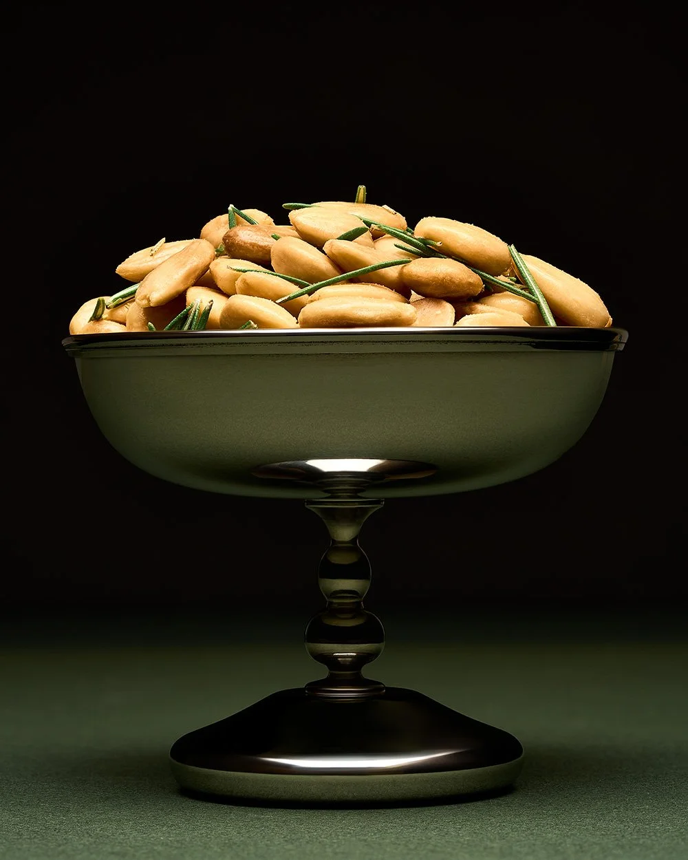 A green decorative bowl filled with pine nuts and sprigs of rosemary, set against a dark background.