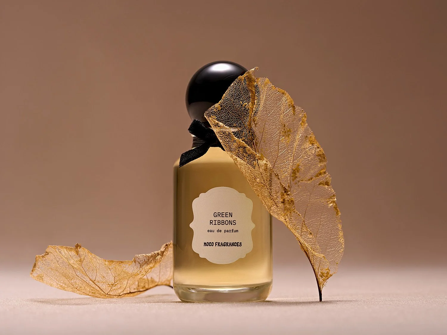 MOCO Green Ribbons perfume bottle surrounded by delicate golden skeletonized leaves against a warm neutral background. Botanical fragrance photography by photographer Sara Anderson.