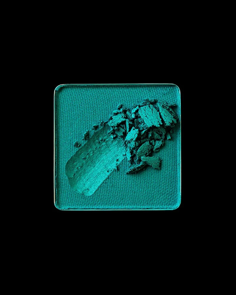 A teal eyeshadow swatch on a square, mesh-textured eyeshadow pan, with a chunk broken off and scattered around, on a black background.