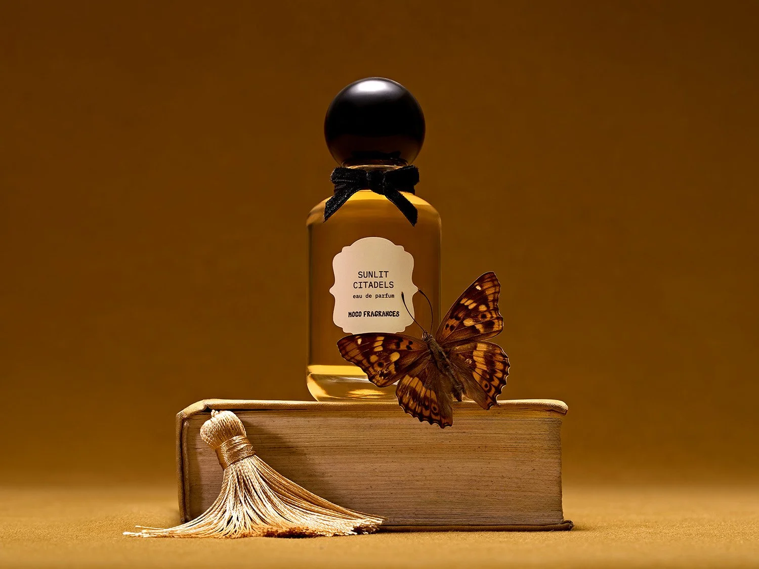 MOCO Sunlit Citadels perfume bottle resting on an antique book with a butterfly and cream tassel. Editorial still-life photography by photographer Sara Anderson.