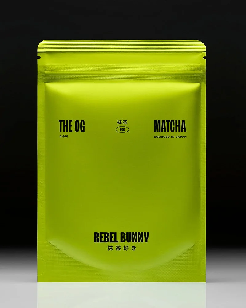 Bright yellow-green packaging for matcha green tea powder with black text that says "The OG Matcha," "Rebel Bunny," and other details, against a black background.