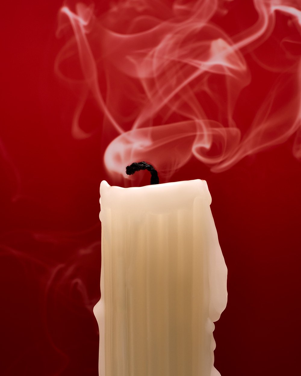 A burning white candle with smoke rising against a red background.