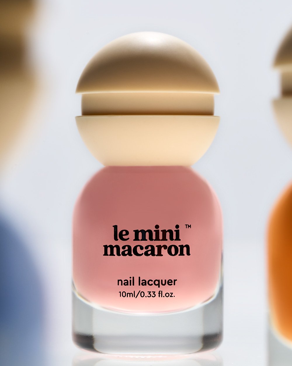A bottle of le mini macaron nail lacquer in pink color, with a beige cap, displayed on a reflective surface with other nail polish bottles partially visible.