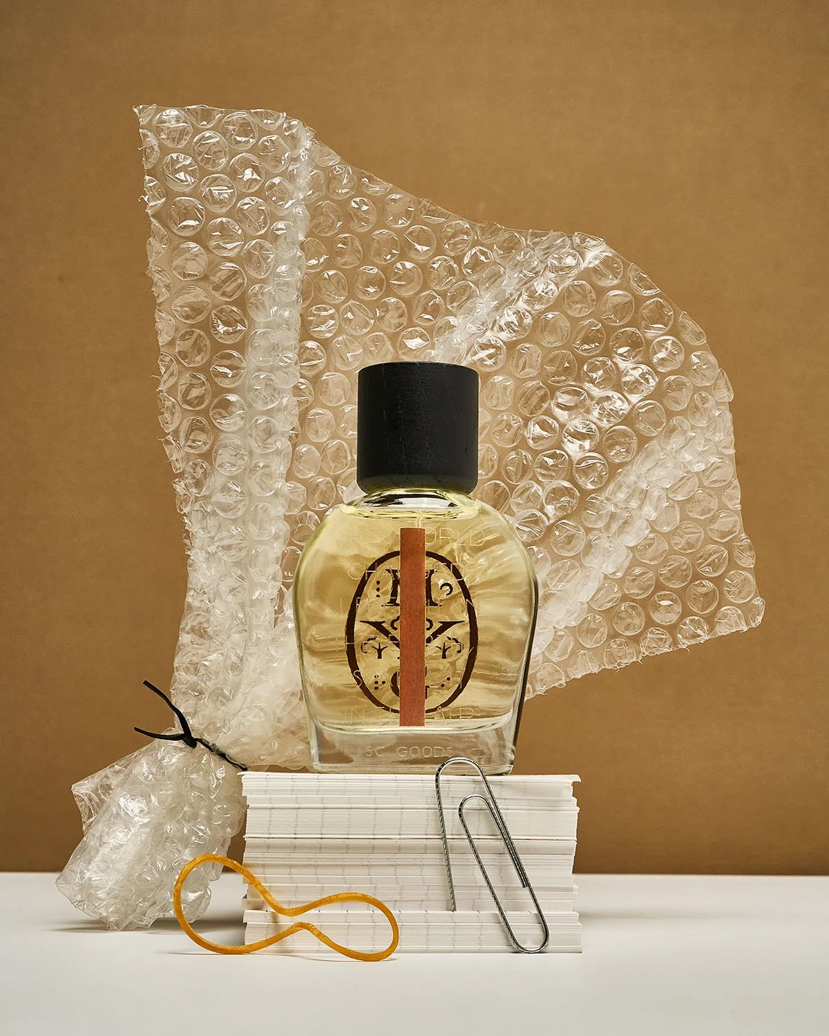 Glass perfume bottle with black cap and gold label displayed on stacked papers, surrounded by bubble wrap, paper clips, and a rubber band.