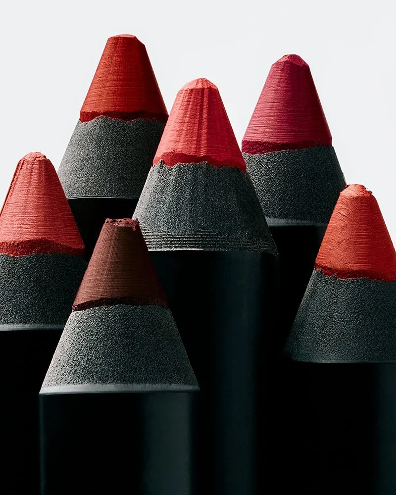 Close-up of seven black and red colored lipstick pencils standing upright against a white background.