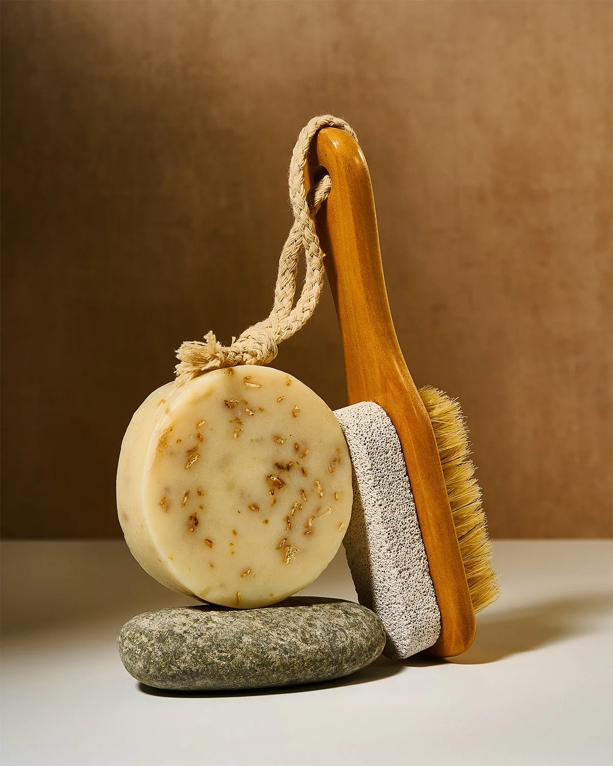 Natural soap, wooden body brush, pumice stone, and hanging rope arranged against a warm backdrop. Wellness product photography by photographer Sara Anderson.