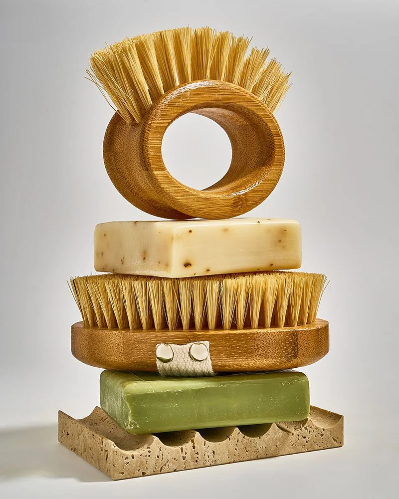 Stacked eco-friendly cleaning tools including a round wooden scrubber, bar soap, brush with bristles, another scrubber, a green soap bar, and a stone base.