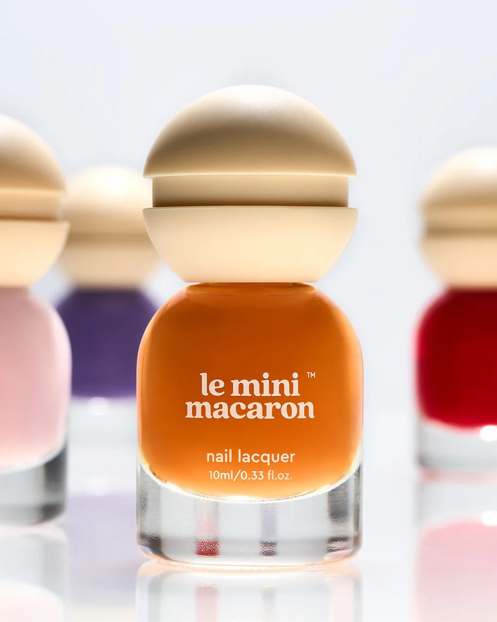 Close-up of a bottle of le mini macaroni nail lacquer on a reflective surface with colorful blurred bottles in the background.