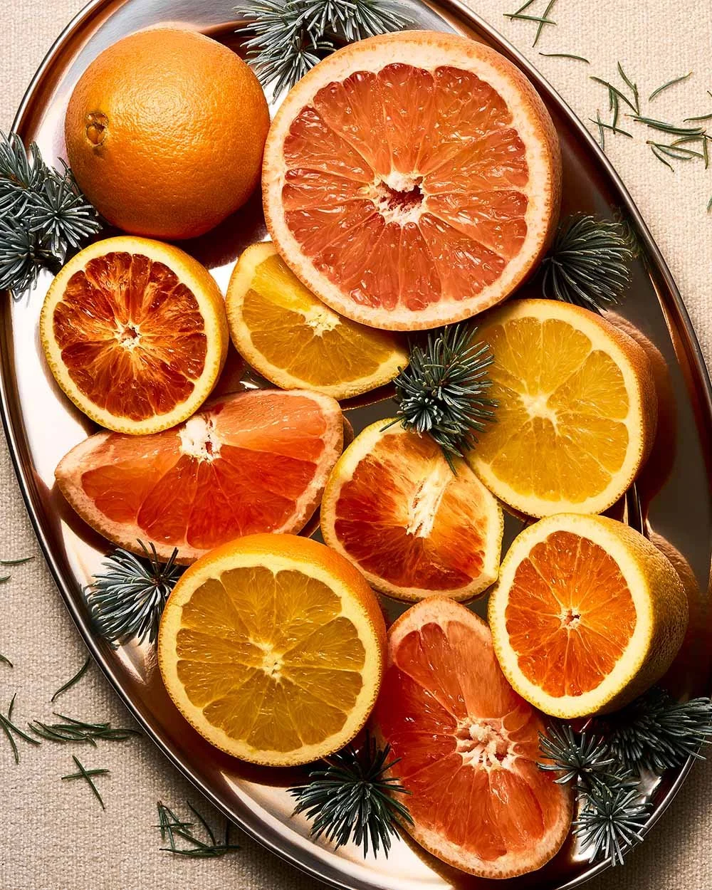 A silver tray with whole and sliced oranges and grapefruits, along with some green pine sprigs, on a light-colored surface.