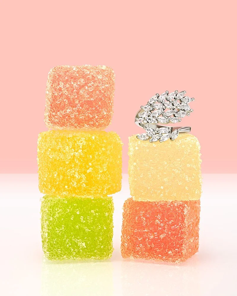 Six colorful sugar-coated gummy candies arranged in two vertical stacks on a pink and white background, with a silver ring adorned with multiple small diamonds placed on top of the right stack.