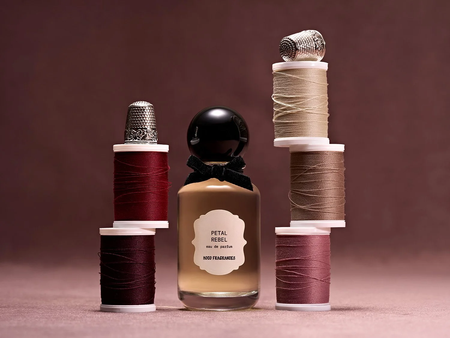 MOCO Petal Rebel perfume bottle framed by burgundy, pink, and cream thread spools topped with silver thimbles. Conceptual fragrance photography by photographer Sara Anderson.
