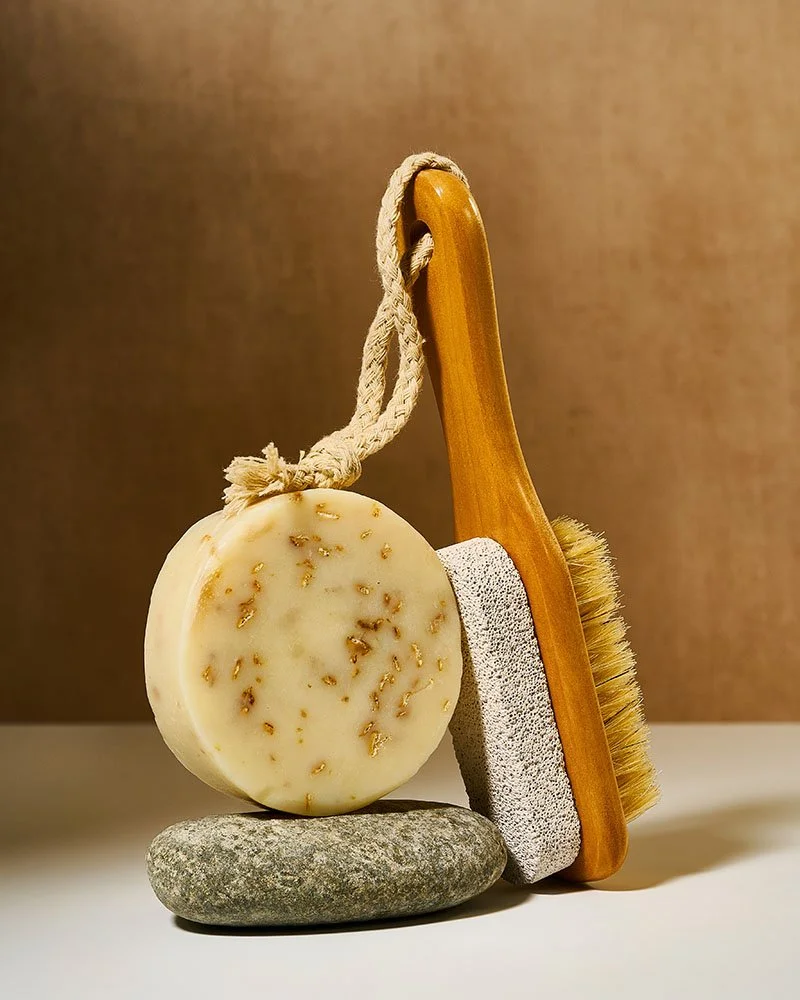 A natural soap with herbs, a pumice stone, a wooden back scrub brush, and a stone all arranged on a white surface with a brown background.