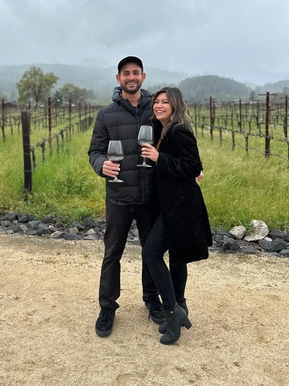 A woman and a man in front of a field holding wine glasses and smiling together